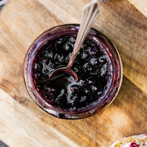 A close-up shot of blueberry jam in a jar with toast on a wooden board.