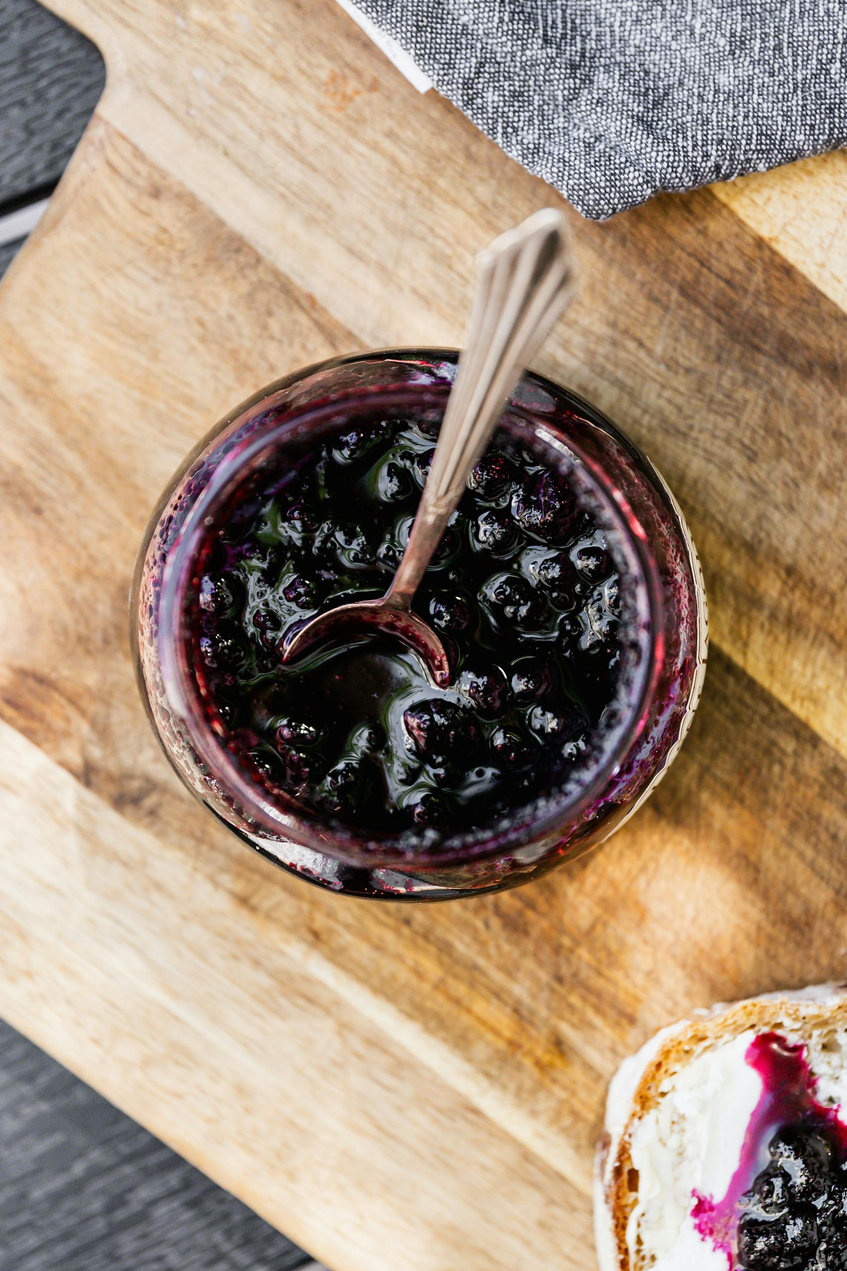 A close-up shot of blueberry jam in a jar with toast on a wooden board.